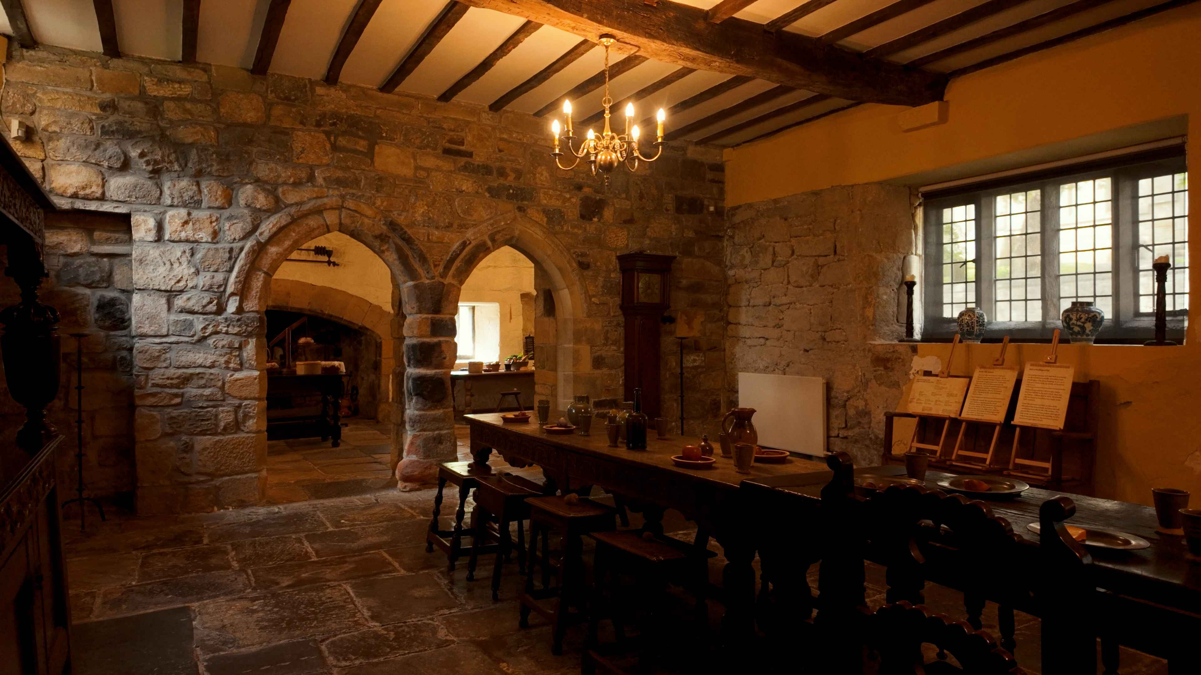 A historic stone-walled dining room with a long wooden table, arched doorways, an old clock, and a lit chandelier, illuminated by natural and artificial light