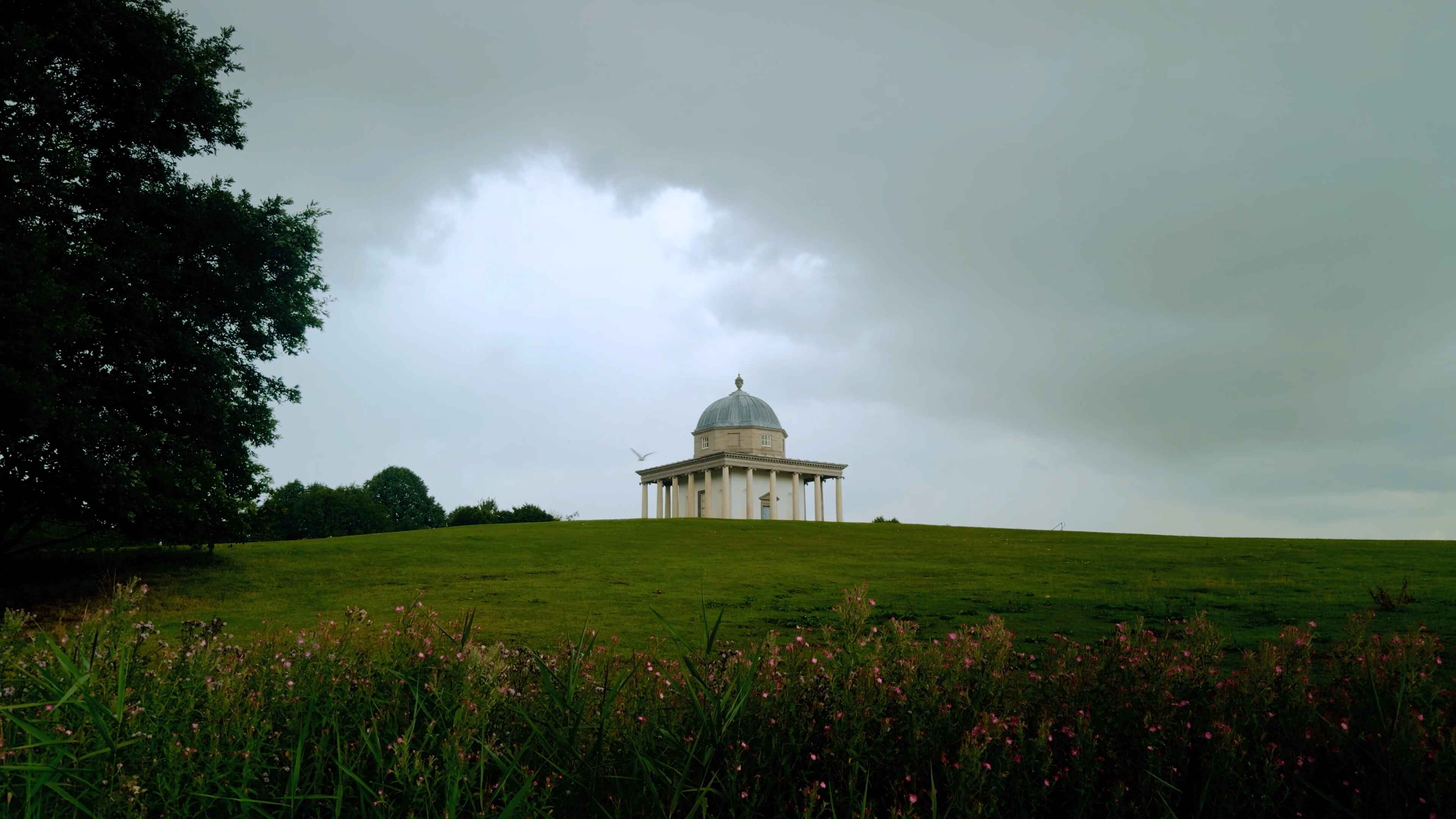 A temple with overcast skies and a bird panning across from the right with flowery foliage in the foreground