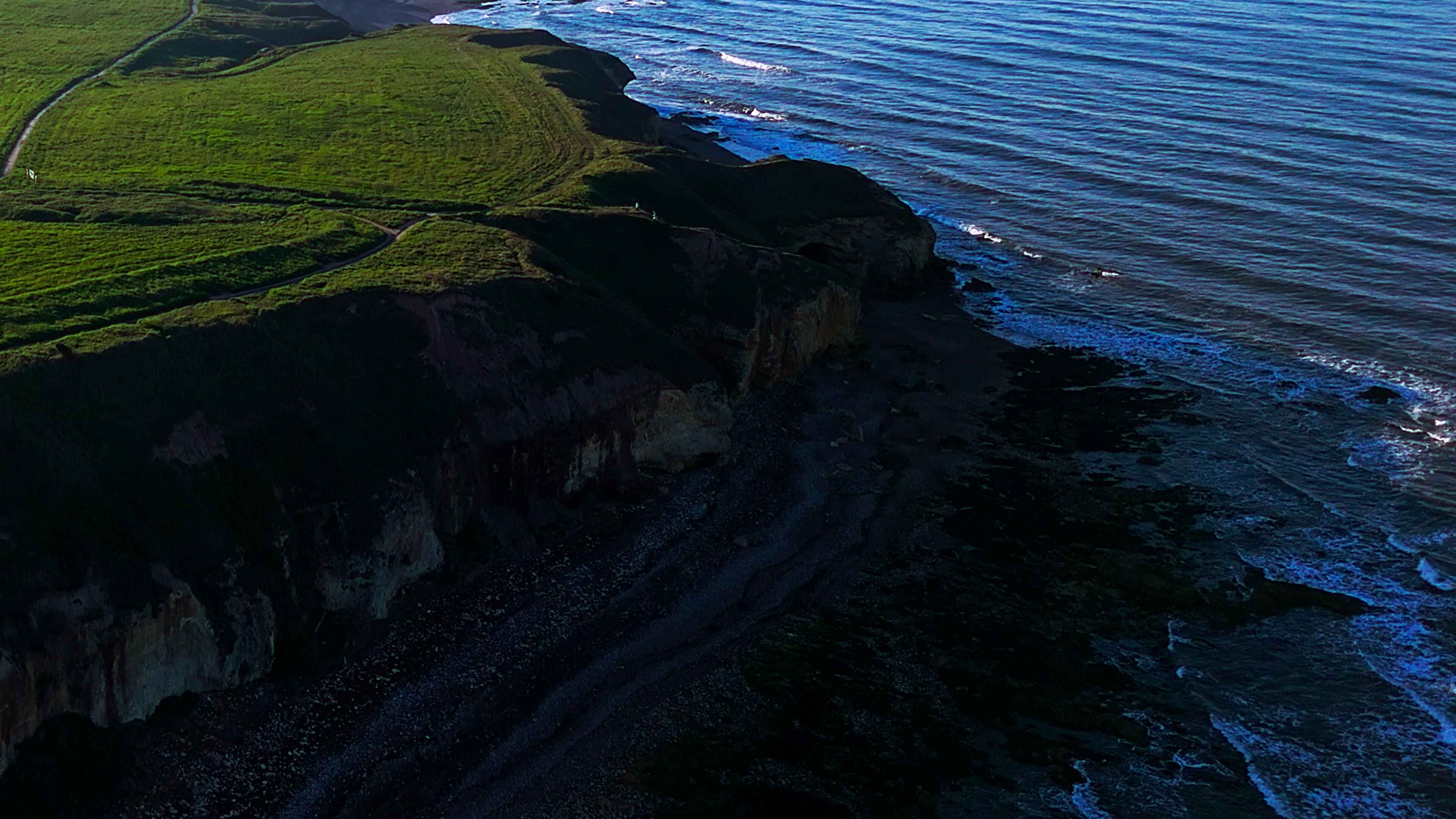 Lush green cliffs to the left overlook a large body of striking blue beach-front water