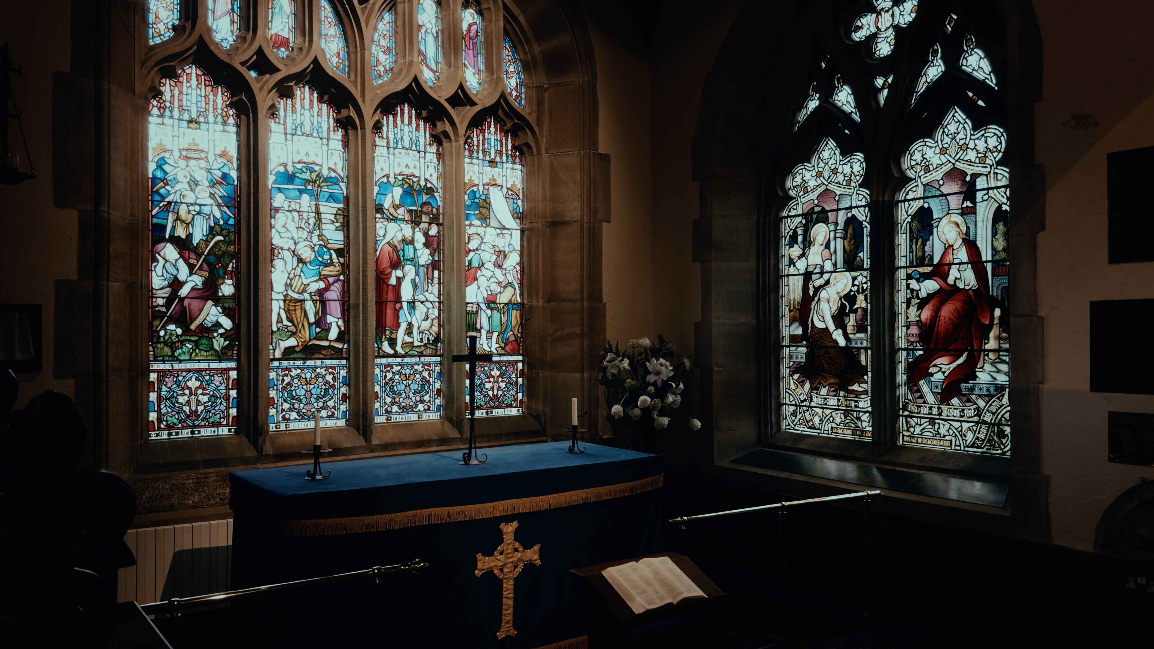 Two stained glass windows meeting at the corner of a church interior with a prayer tabletop and crosses centred above